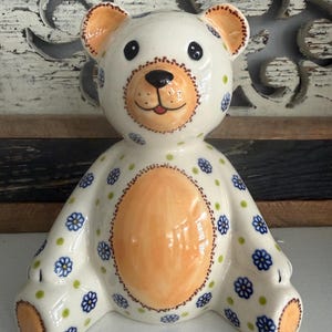 May include: A ceramic bear figurine featuring a cream-colored base with a hand-painted pattern of small blue flowers and green dots. The bear has a light orange belly, ear interiors, and foot pads, with a brown stitched-style border detail.