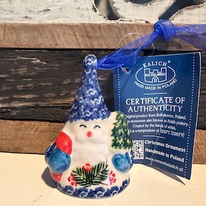 May include: A ceramic bell-shaped ornament featuring a gnome with a blue patterned hat, holding a green tree and a red gift. The white base displays a floral motif. A blue ribbon is attached to the top, alongside a tag labeled CERTIFICATE OF AUTHENTICITY.