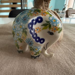 May include: A ceramic elephant figurine featuring a white base with a colorful floral pattern in shades of blue, orange, and green. The elephant has a dark blue circular design on its ear and a painted eye with visible eyelashes.