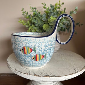 May include: A ceramic mug with a unique handle design. The mug features a blue and white wave pattern with two colorful fish illustrations. The handle and rim are trimmed in dark blue. The mug is displayed on a white, distressed wooden stand.