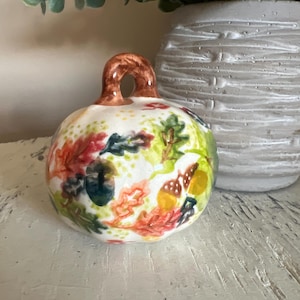 May include: A ceramic pumpkin-shaped decorative item with a brown stem and a white base. The pumpkin is adorned with colorful autumn leaves and acorns in shades of red, orange, green, and yellow. The item is placed on a white surface.