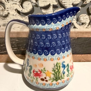 May include: A ceramic pitcher featuring a dark blue scalloped pattern along the top rim and a cream-colored base decorated with colorful illustrations of red crabs, yellow starfish, green seaweed, and blue seahorses around the body.