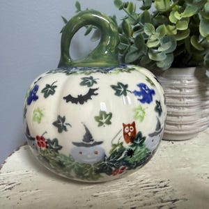 May include: A ceramic pumpkin figurine with a glossy cream-colored surface featuring hand-painted motifs of black bats, blue ghosts, orange owls, green ivy leaves, and small grey figures wearing pointed hats, topped with a green glazed stem.
