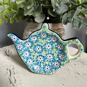 May include: A ceramic teapot-shaped tea bag holder with a blue floral pattern. The holder has a dark blue rim and handle, and is decorated with white and blue flowers on a light blue background. The holder is on a white surface.