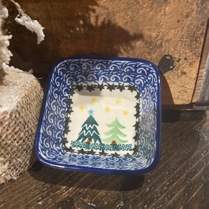 May include: A small blue and white ceramic dish with a square shape and a design of two green Christmas trees with yellow stars. The dish has a blue and white swirl pattern around the edge.
