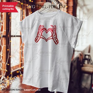 May include: A white t-shirt featuring a red skeleton hand heart design. The design depicts two skeleton hands forming a heart shape.