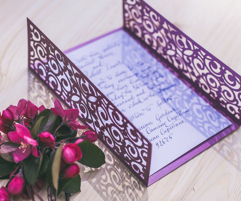 May include: Purple wedding invitation with a laser cut design and a white inner card with calligraphy. The invitation is on a wooden surface with pink flowers.