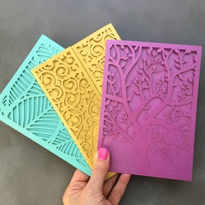 May include: Three laser cut cards with intricate designs. The card on the left is teal blue with a leaf pattern. The middle card is yellow with a floral pattern. The card on the right is purple with a tree branch pattern.