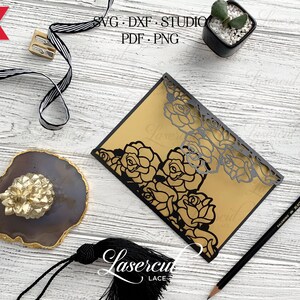 May include: A black and gold laser cut wedding invitation with a floral design. The invitation is on a wooden surface with a pencil, a pencil sharpener, and a small potted plant.