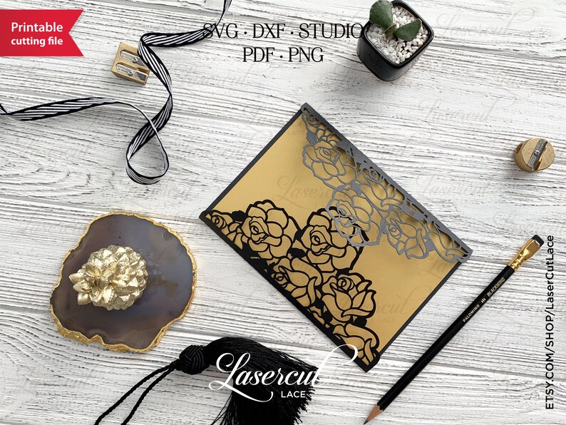 May include: A black and gold laser cut wedding invitation with a floral design. The invitation is on a wooden surface with a pencil, a pencil sharpener, and a small potted plant.