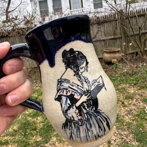 May include: A ceramic mug with a dark blue rim and handle. The mug features a hand-drawn illustration of a woman in a dress reading a book. The mug is beige with blue accents and a black illustration.