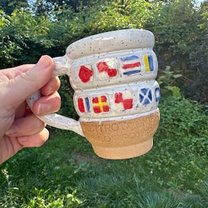May include: A handmade ceramic mug with a speckled white glaze and a tan base. The mug features colorful flag designs around the body and the text "TROT UNIFORM" on the base. The handle is curved and matches the speckled glaze.