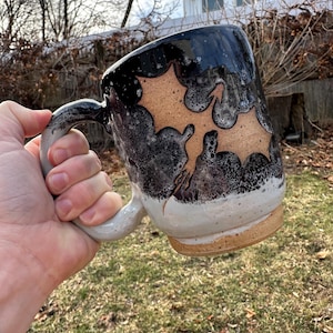 May include: A black and white ceramic mug with a brown glaze and a design of two holly leaves. The mug has a speckled glaze and a handle.