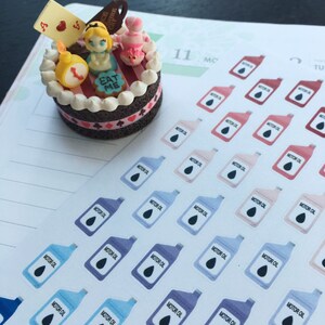 May include: A miniature cake decorated with Alice in Wonderland themed figurines, including Alice, the Mad Hatter, and a playing card. The cake is decorated with red and white stripes, a chocolate base, and the words "Eat Me".