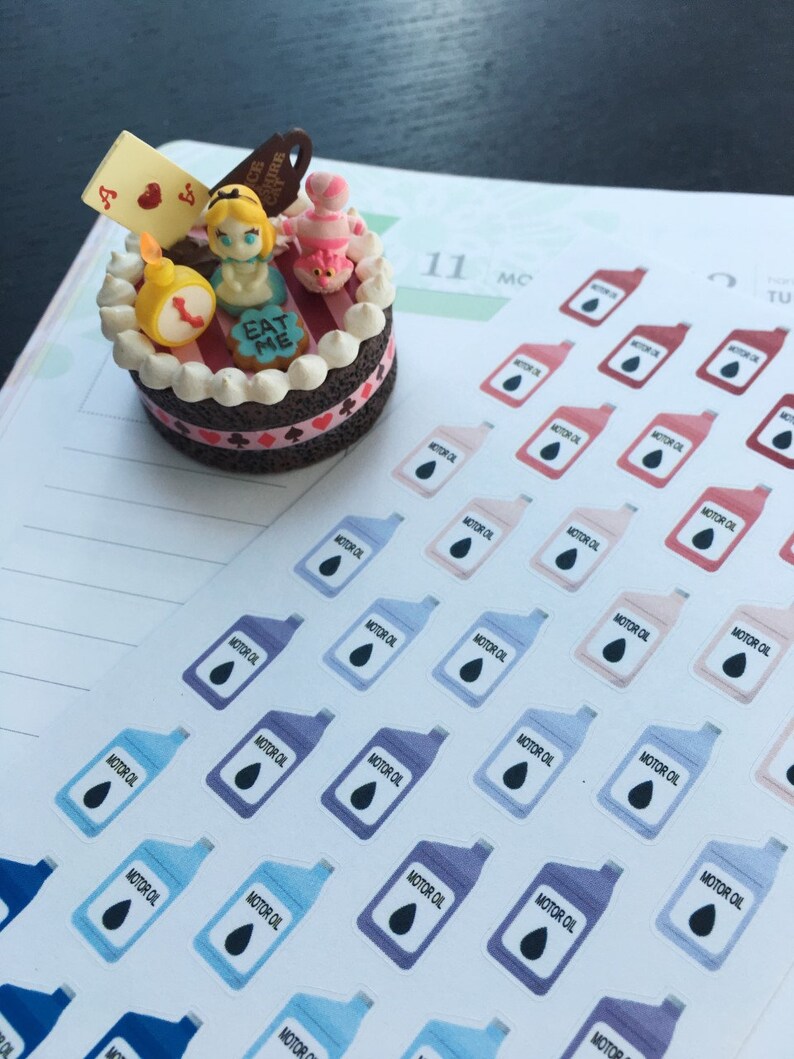 May include: A miniature cake decorated with Alice in Wonderland themed figurines, including Alice, the Mad Hatter, and a playing card. The cake is decorated with red and white stripes, a chocolate base, and the words "Eat Me".