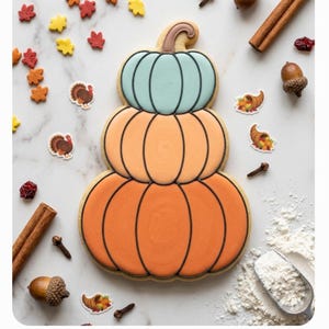 May include: A stacked pumpkin-shaped sugar cookie with three tiers in light blue, peach, and orange icing, decorated with black outlines. The cookie is surrounded by small autumn-themed sprinkles, cinnamon sticks, acorns, cloves, and a metal scoop of flour.