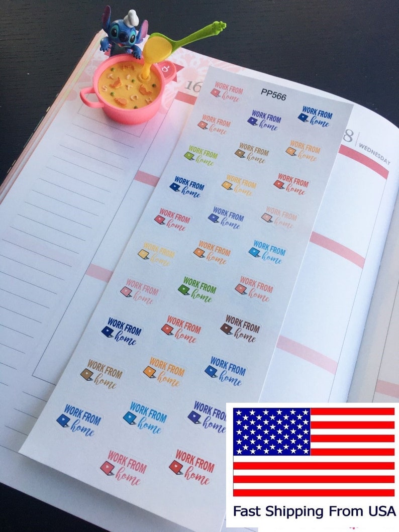 May include: A planner is open to a double-page spread with stickers that say "WORK FROM home" in various colors. A small figurine of a blue character with a spoon is near a pink bowl. The American flag is in the bottom right.