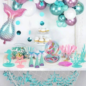 May include: A mermaid-themed party table setting with teal and pink decorations. The table features a variety of items, including plates, cups, napkins, and centrepieces. The centrepieces include a mermaid tail, a starfish, and a seahorse. The table is decorated with a teal and white net.