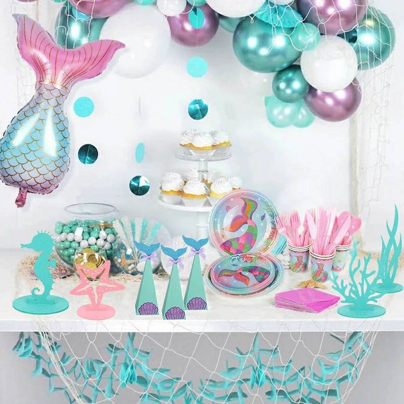 May include: A mermaid-themed party table setting with teal and pink decorations. The table features a variety of items, including plates, cups, napkins, and centrepieces. The centrepieces include a mermaid tail, a starfish, and a seahorse. The table is decorated with a teal and white net.