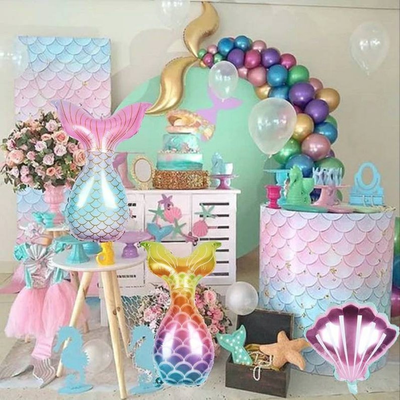 May include: A mermaid-themed party decoration set featuring a gold foil mermaid tail, iridescent balloons, and a pink and blue shell-shaped balloon. The set also includes a pink and blue mermaid tail balloon and a pink and purple mermaid tail balloon.