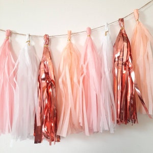 Puede incluir: Una guirnalda de borlas de papel de seda en tonos rosa, melocot&oacute;n y blanco con una borla de oro rosa met&aacute;lico. Las borlas est&aacute;n enhebradas en una cuerda de yute.