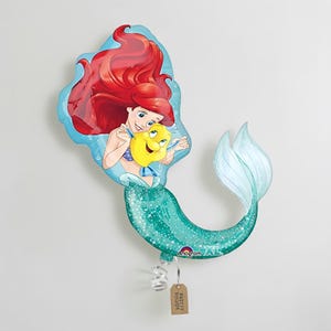 May include: Ariel the Little Mermaid foil balloon. The balloon features Ariel with red hair and a yellow fish. The mermaid's tail is teal with glitter. The balloon is attached to a tag that says "Party Junior".