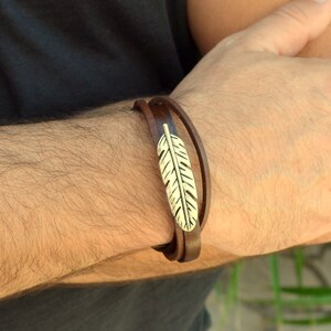 Könnte beinhalten: Ein braunes Lederarmband mit einem silbernen Federschmuck. Das Armband ist um das Handgelenk einer Person gewickelt.