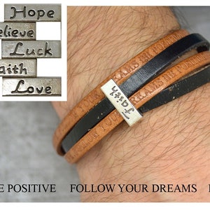 Op de afbeelding: Een bruin en zwart leren armband met een zilveren bedel waarop "Hard Work" staat. De armband heeft de woorden "Be Positive" en "Follow Your Dreams" gegraveerd.