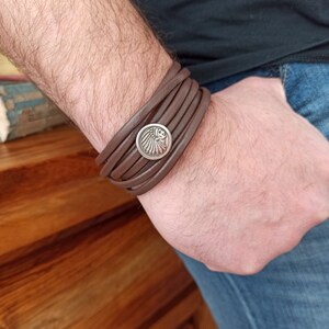 Könnte beinhalten: Ein braunes Lederarmband mit einem silbernen runden Anhänger, der ein indianisches Kopfschmuckdesign zeigt.