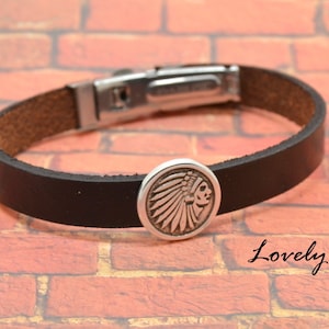 Puede incluir: Un brazalete de cuero negro con un cierre plateado y un dije plateado con un diseño de tocado nativo americano.