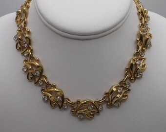 May include: A gold tone necklace with a delicate design featuring small clear rhinestones. The necklace is made of a chain with a series of small, leaf-shaped links.