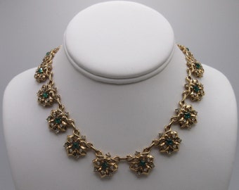May include: Gold-toned necklace featuring floral designs with emerald green and clear crystal accents. The necklace is displayed on a white bust, showcasing its intricate details and elegant design. A classic piece of jewelry.