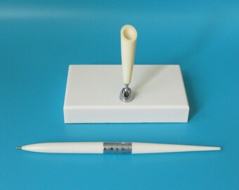 May include: White pen holder with a white pen on a blue background. The pen holder has a white base and a white pen holder with a silver top. The pen is white with a silver band.
