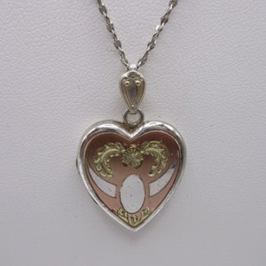 May include: A silver heart-shaped locket with a gold-toned design. The locket has a white oval in the center and is on a silver chain.