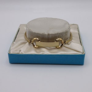 May include: A gold-toned expandable bracelet with a rectangular nameplate and two circular links. The bracelet is displayed in a blue box with white satin lining.