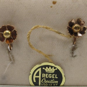 May include: A pair of gold flower-shaped earrings with a small gold bead in the center of each flower. The earrings are on a white background with a tag that reads "A Regel Creation 1/20-12 KT".