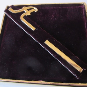 May include: A gold-coloured tie clip with an initial design, presented on a dark purple velvet insert within a gold-edged box. The clip features a stylized letter and a rectangular bar.