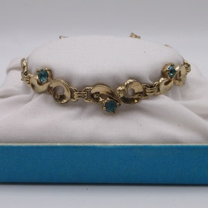 May include: A gold bracelet with a delicate, ornate design featuring blue gemstones. The bracelet is displayed on a white velvet cushion inside a blue box.