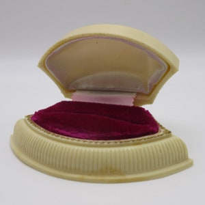 May include: A white plastic ring box with a pink velvet lining and a gold trim. The box is open and the velvet lining is visible.