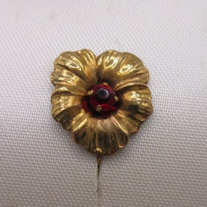 May include: A gold-toned flower pin with detailed petals and a central red gemstone. The pin is designed in the shape of a flower, with the petals radiating outwards from the center. The red gemstone adds a pop of color.