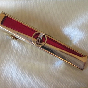 May include: A gold tie bar with a red enamel inlay and a circular emblem featuring the letters "LFT" in blue, white, and red.