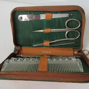 May include: Open brown leatherette travel manicure kit with a green interior. The kit contains a nail file, scissors, tweezers, and a comb. The kit is labeled "PIGSKIN" on the leather strap.