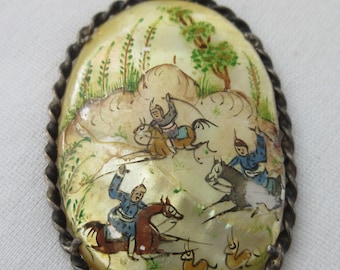 May include: A silver pendant with a hand-painted scene of people riding horses in a landscape. The pendant is made of mother-of-pearl and has a twisted silver border.