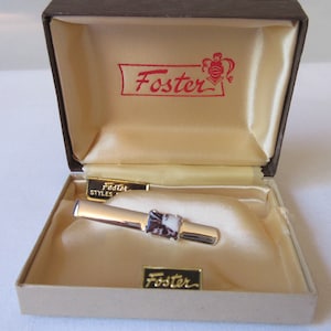 May include: A silver tie bar with a brown and white stone in a beige box with the word "Foster" printed in gold lettering.