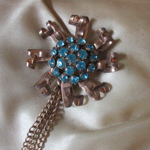 May include: A vintage brooch with a large cluster of turquoise blue rhinestones in the center. The brooch is made of gold-toned metal and has a long chain attached to it.