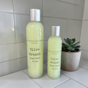May include: Two bottles of Olive Branch Body Wash by Tipsy Goat Soap Company. The bottles are filled with a pale green liquid and have silver caps. The larger bottle is 8.0 oz, and the smaller is 4.0 oz. A small potted succulent is to the right.