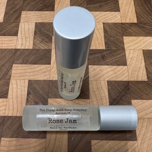 May include: Two silver roll-on perfume bottles with a clear liquid inside. The label on the bottle reads "The Tipsy Goat Soap Company, Warsaw, IN, Rose Jam, Roll On Perfume, 10mL."