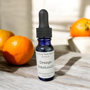 May include: A small blue glass bottle with a black dropper top. The bottle is labeled "The Tipsy Goat Soap Co. Warsaw, IN" and "Orange Patchouli Car Freshener Oil 0.5oz (14g)".