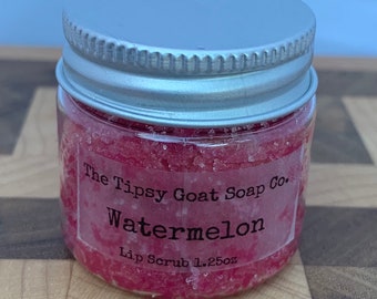 Op de afbeelding: Een klein glazen potje met een zilveren deksel gevuld met roze lippen scrub. Het etiket vermeldt "The Tipsy Goat Soap Co. Watermelon Lip Scrub 1.25oz."
