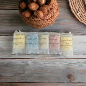 May include: Four small, clear plastic tubes containing body butter sticks. The tubes are labeled with the names "Honeysuckle", "Temptation", "Apple Cider", "Rose Jam", and "Lemon Pound Cake". Each tube has a small sample of body butter inside.